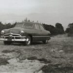 A black and white photo of an old car on a dirt road, exuding vintage charm.
