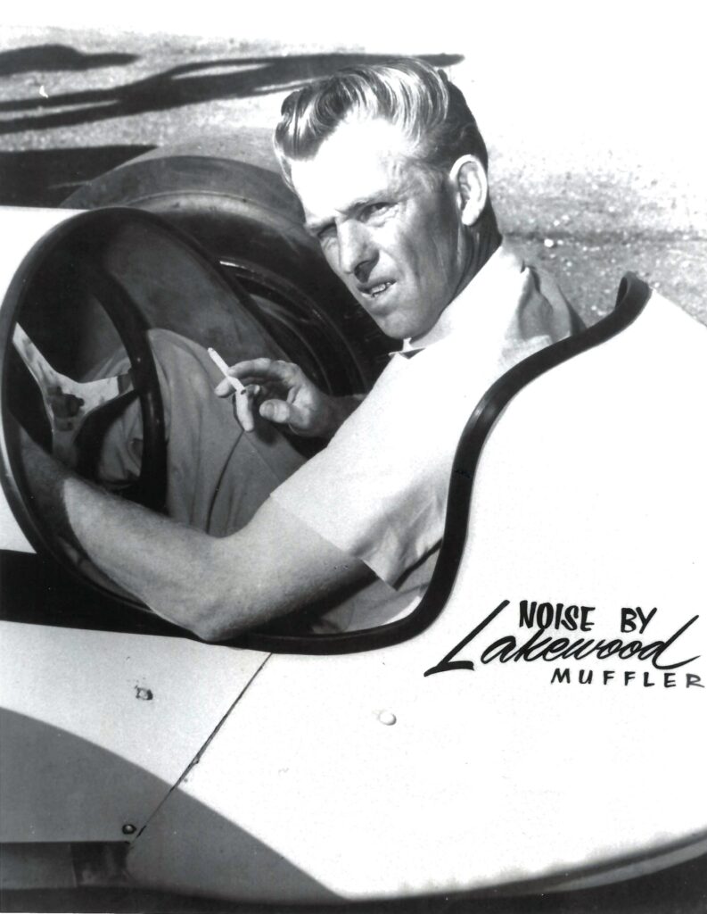 A black and white photo of a man in a race car speeding through a drag strip.