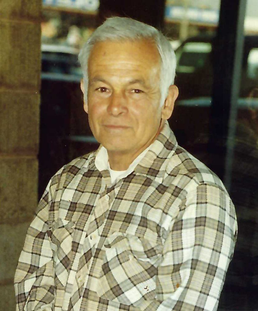 An older man in a plaid shirt standing beside a drag racing building.