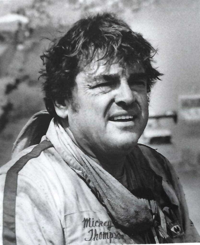 A black and white photo of a man in a jacket, standing near a drag racing car.