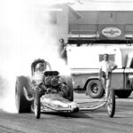 An adrenaline-fueled man races a drag car with smoke billowing behind him.