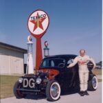 A man standing next to a hot rod car in front of a Drag Racing sign.