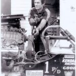 An old photo of a man standing next to a drag race car.