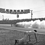 An old photo of a man drag racing on a track with smoke coming out of his car.