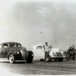 An old black and white photo of a man drag racing on a motorcycle next to a group of cars.