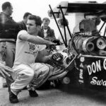 A man kneeling down next to a race car in a drag racing competition.