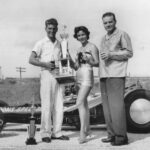 A man and woman triumphantly stand next to a trophy-winning drag racing car.