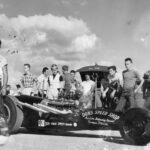 A group of people enthusiastically gathered around a drag racing car.
