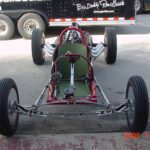 A red and silver race car parked in front of a truck at a drag racing event.