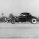 A black and white photo of an old car seen in a drag racing scene.