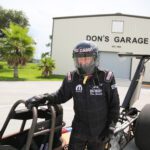A man in racing gear standing next to a drag race car in front of a garage.
