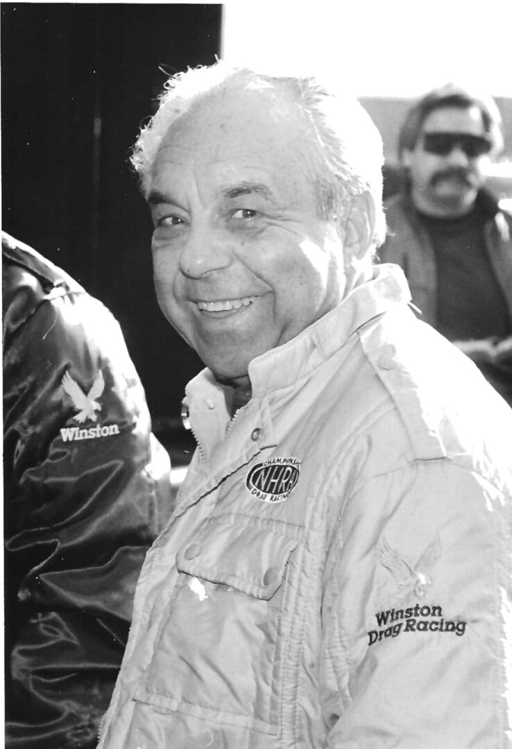 A black and white photo of an older man smiling, possibly reminiscing about his drag racing days.