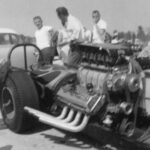A group of men standing around a car at a drag racing event with a big engine.