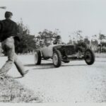 An old black and white photo of a man drag racing in a car.