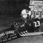 An old photo of a man standing next to a drag racing car.