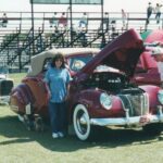 A woman standing next to a red car at a drag racing event.