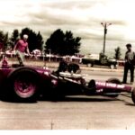 An old photo of a purple drag car. The photo captures the excitement and energy of drag racing as this vibrant purple car speeds down the track, leaving a trail of smoke behind.