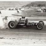 An old photo of a drag race car with smoke billowing out of it.