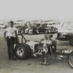 An old photo of a man standing next to a car after a drag racing event.