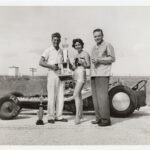 Three people standing next to a drag racing car with trophies.