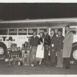 A group of people standing in front of a bus with drag racing trophies.