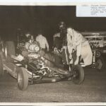 An old photo of a man standing next to a drag car at a drag racing event.