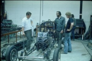 Three men discussing car chassis construction.