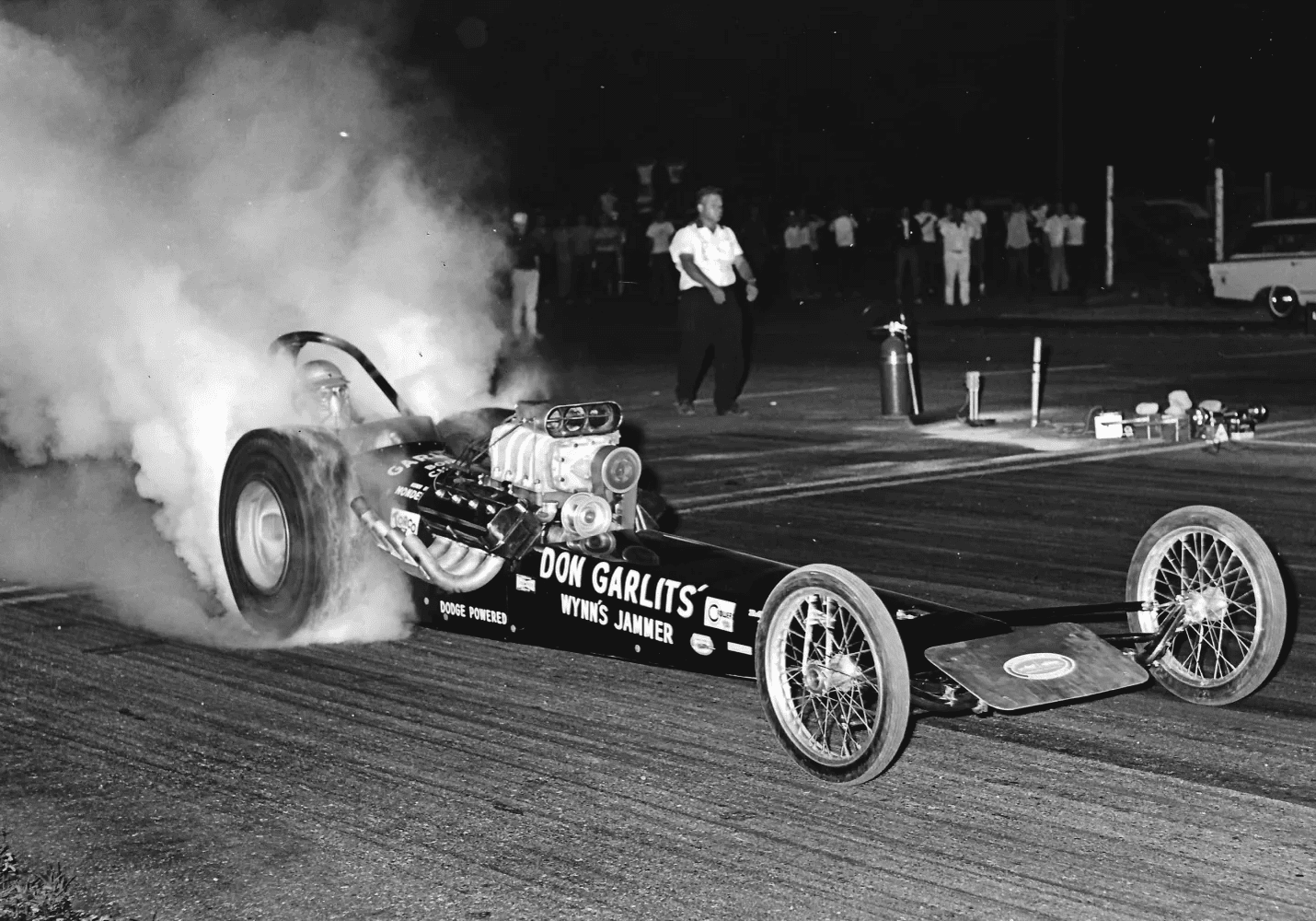 A black and white photo of a drag car with smoke billowing out of it during an intense drag racing event.